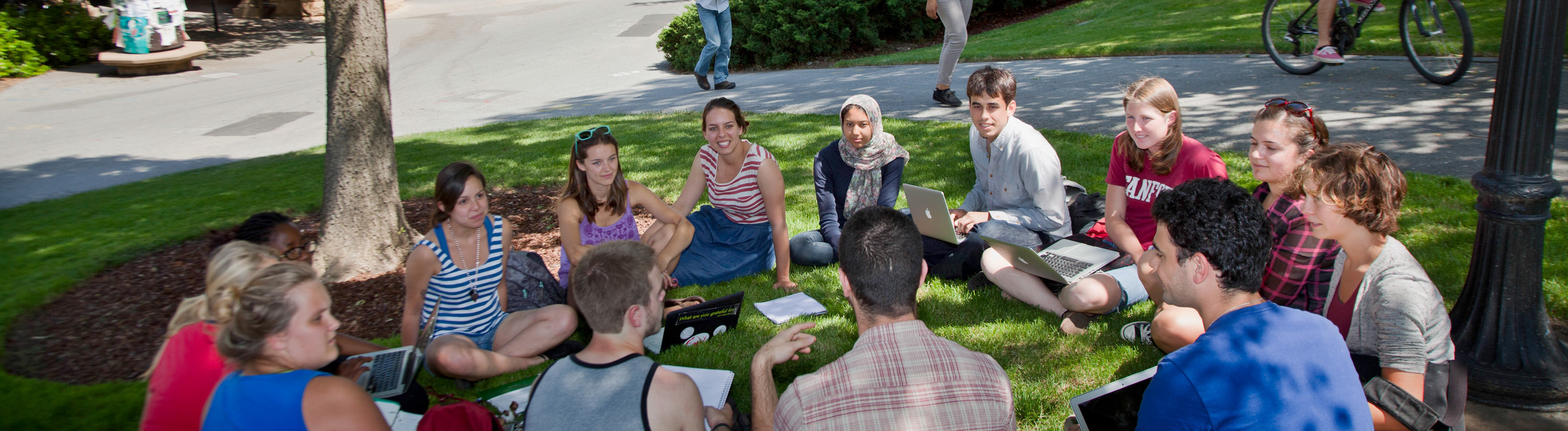 A diverse group of students sit together in a circle on a grassy hill outside the main quad