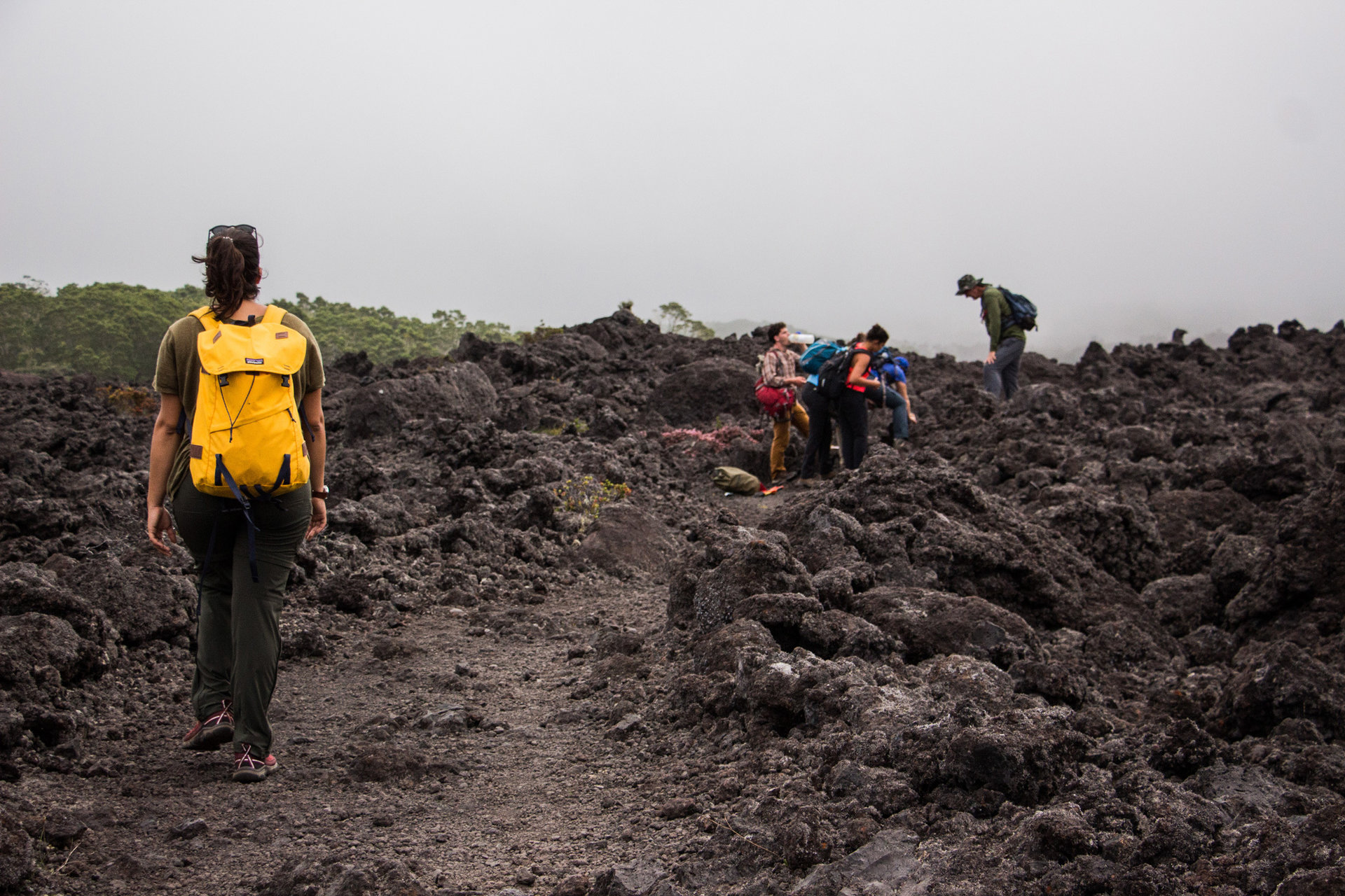 A female student with a bright yellow backpack walks towards the class in a volcanic rock field in Hawaii