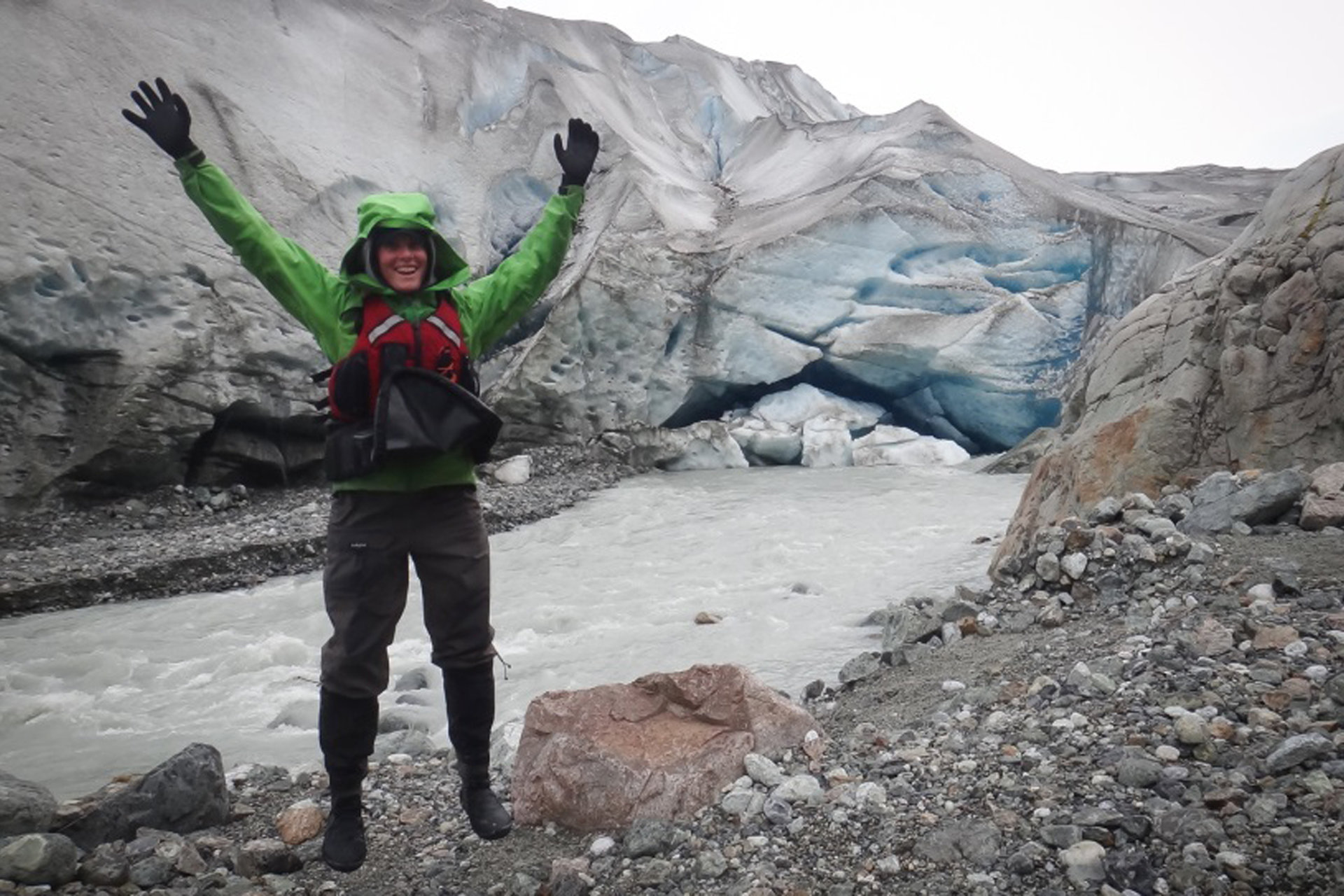 A student lifts her arms up in excitement as she stands near an Alaskan glacier